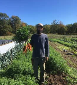 Jeff Benton of Orange Circle Farm Farmer Jeff Benton stand in the pathway between two lush beds of carrots. He is holding a bunch of bright orange carrots by their green tops.