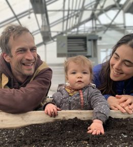 triple-chick-farm Two smiling farmers look on as a baby plays with potting soil