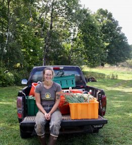 YEA_2024_New Spoke Janelle truck bed A farmer smiles sitting in the back of a truck bed filled with bins of vegetables
